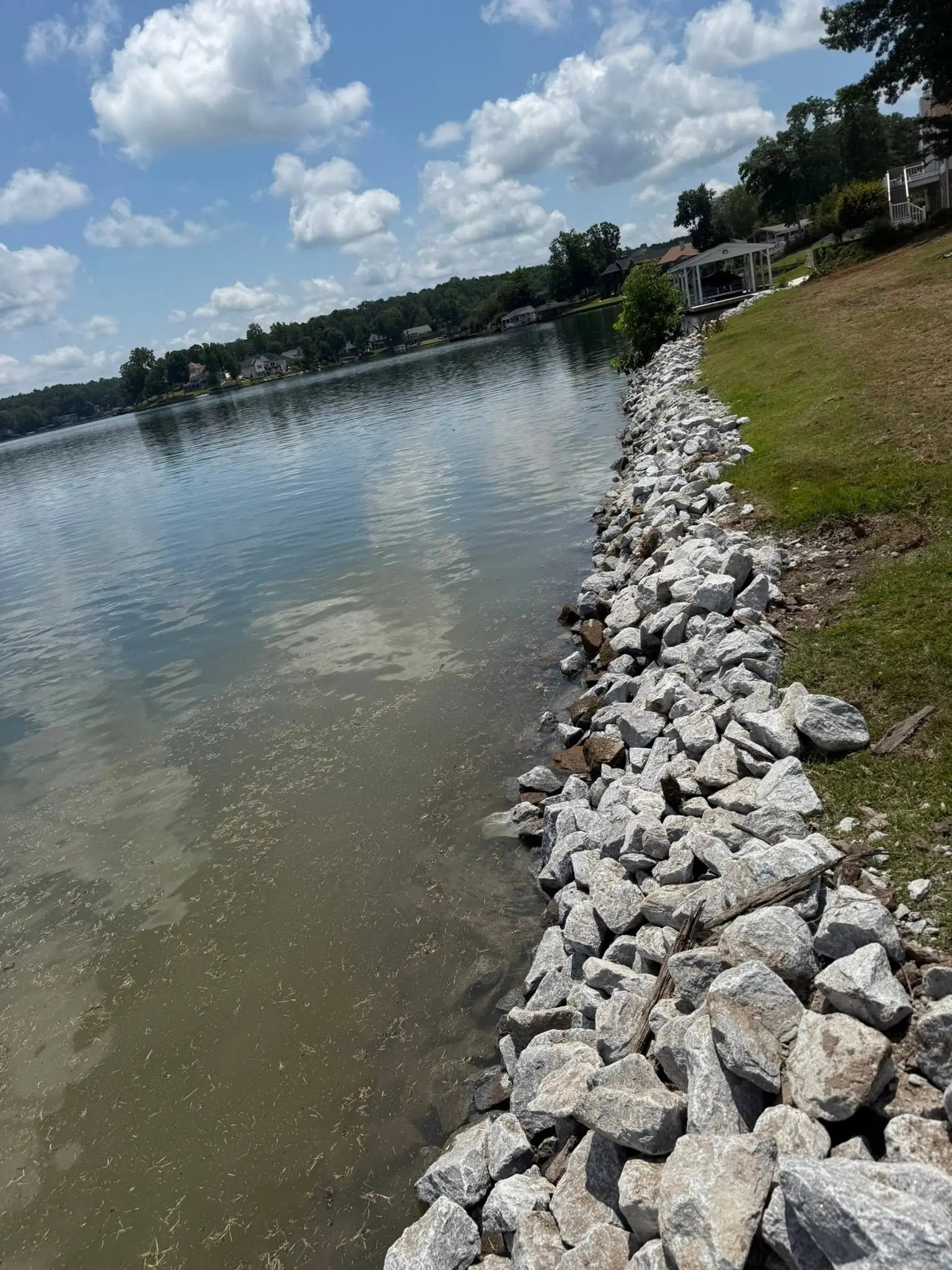 Rip-rap stone seawall along a grassy lake shoreline, lake homes on the far bank