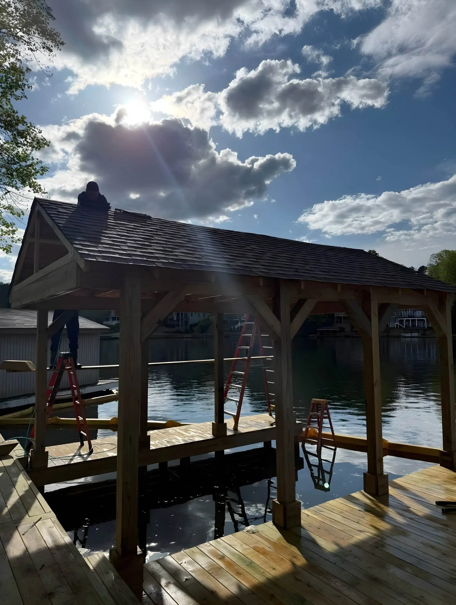 Worker installing shingles on a boathouse gable roof over the water; one gable end still open/unsheathed.
