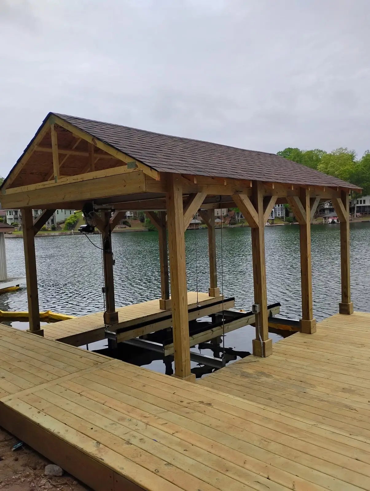 Covered boathouse with boat lift mechanism and a shingled gable roof, lake homes across the water; dock deck is unstained with no rail installed, silt curtain still visible in the water