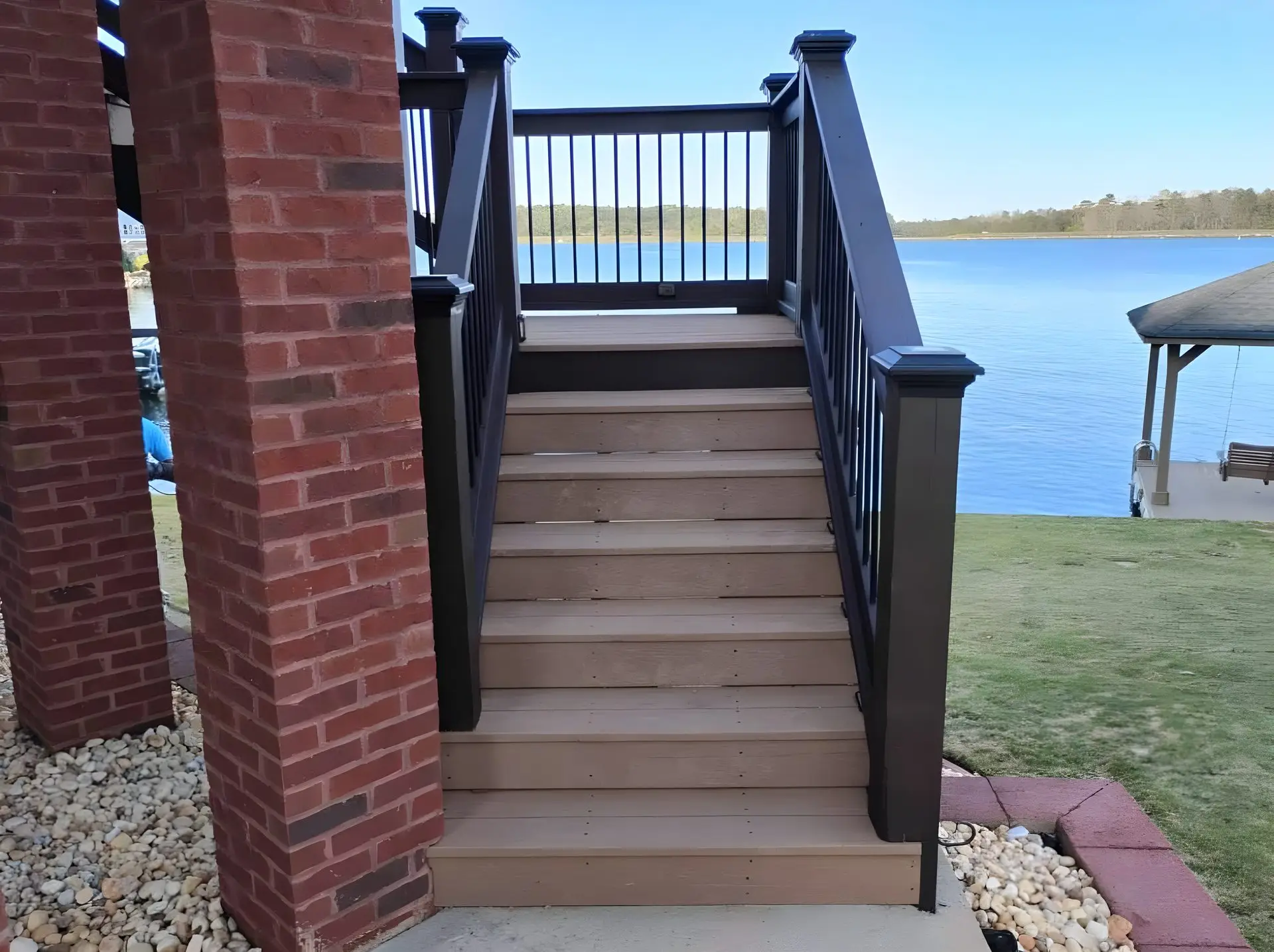 Composite deck stairs with black rail descending between two brick columns to a lakeside patio, open lake visible beyond.