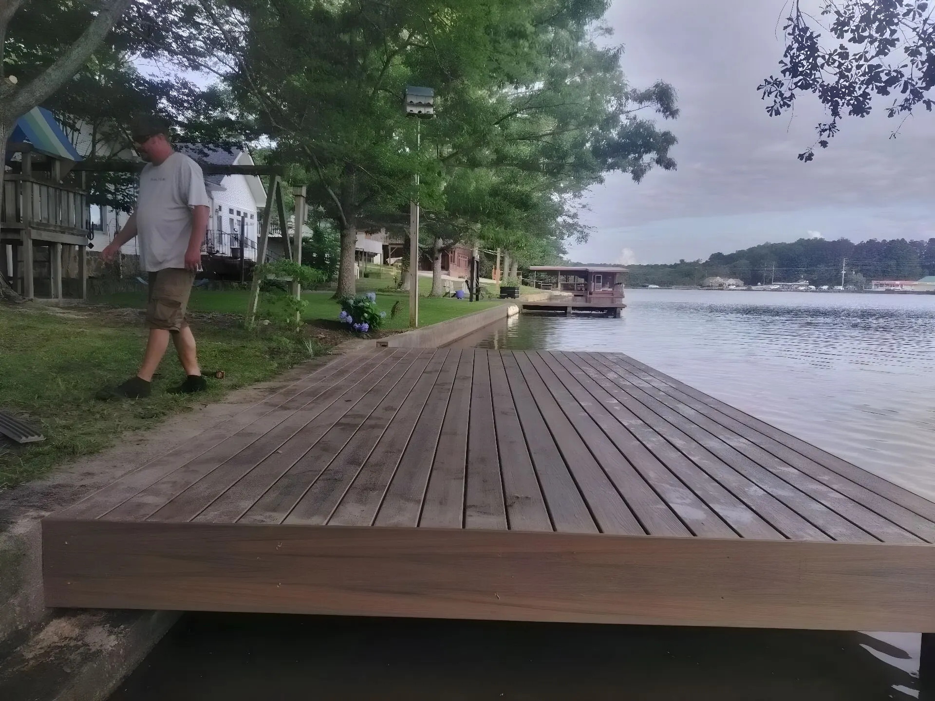 Completed composite dock platform at the water's edge, a man walking the bank beside it, lake homes and open water behind