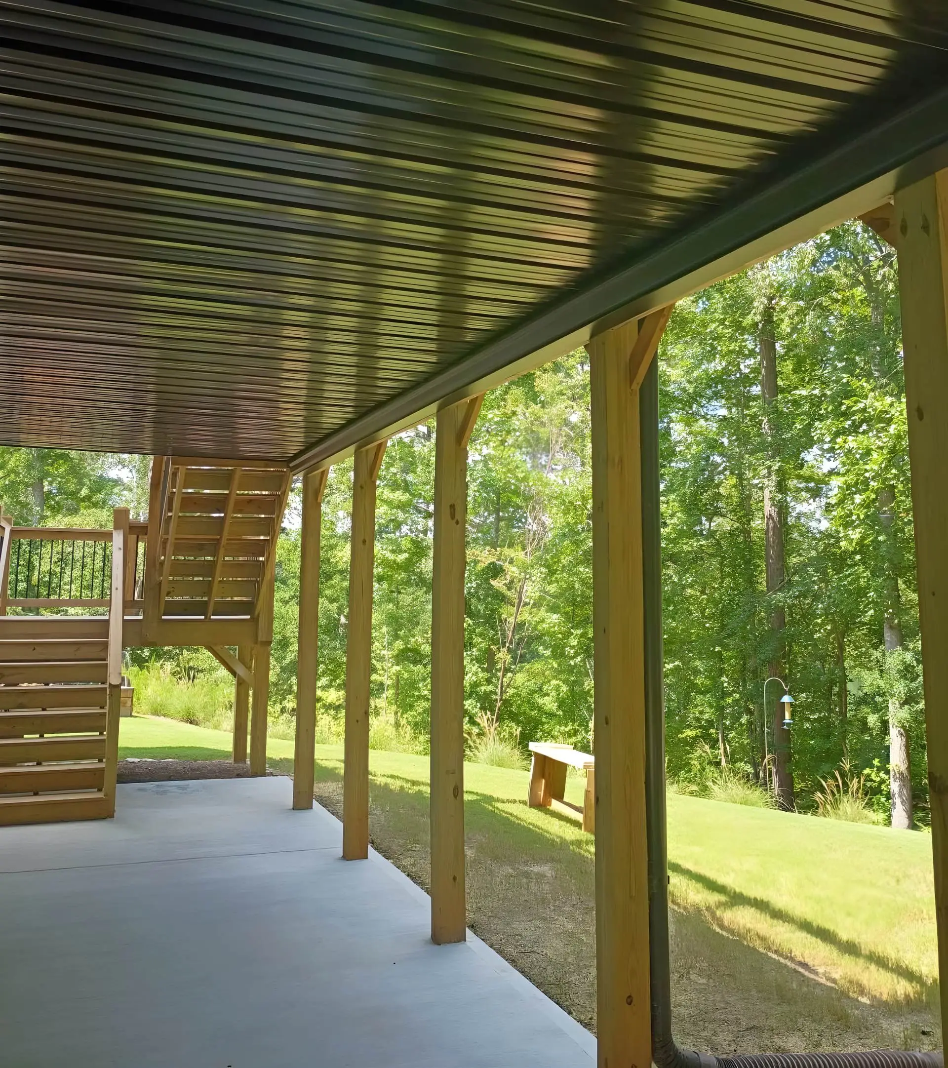 Covered patio with black corrugated metal ceiling, staircase up to the second-level deck, concrete floor, and a freestanding wood bench out on the sunlit lawn beyond the posts.