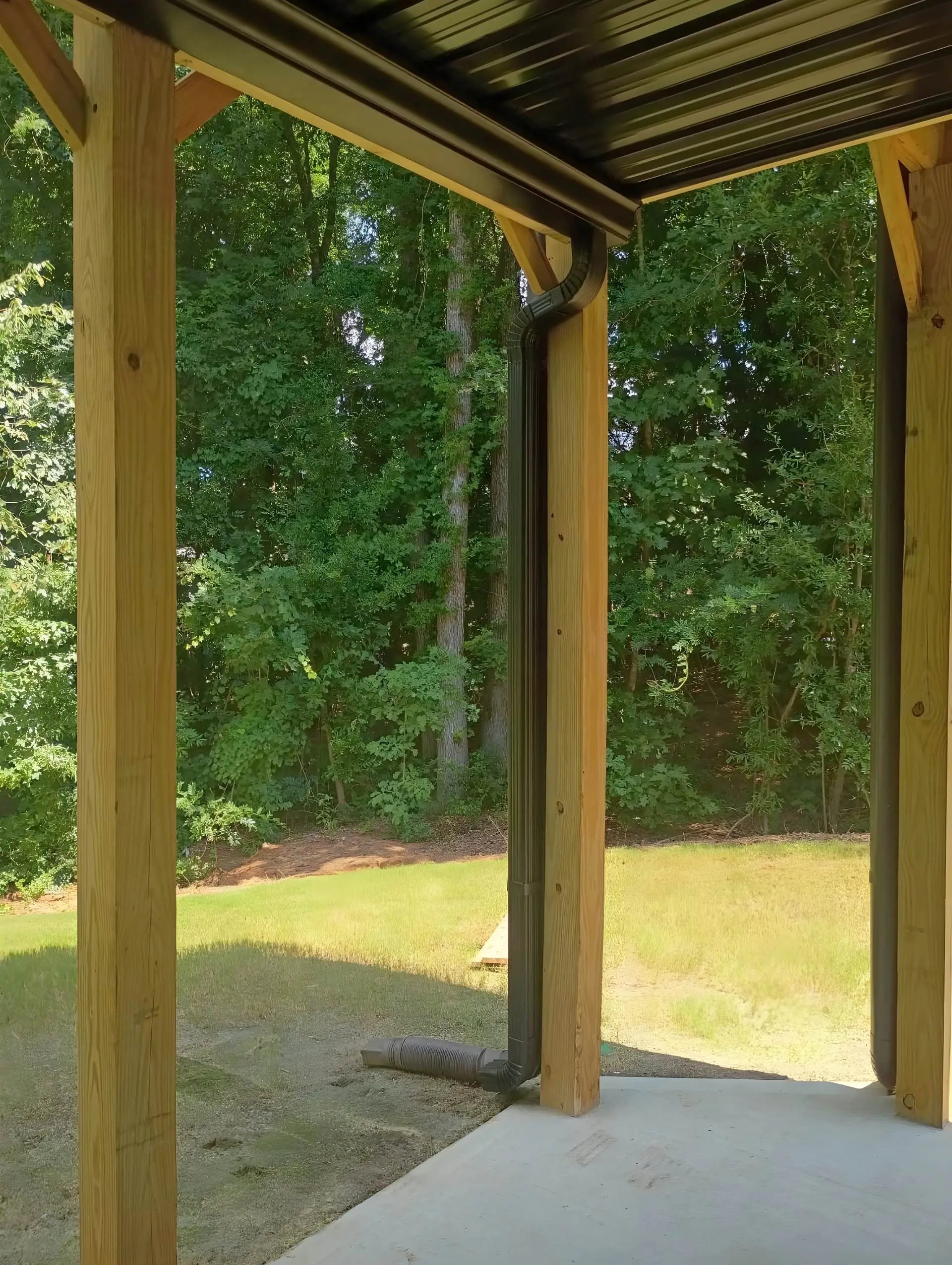 New covered patio with a corrugated metal roof and wood posts, clean poured concrete slab facing a mowed lawn and wooded tree line.