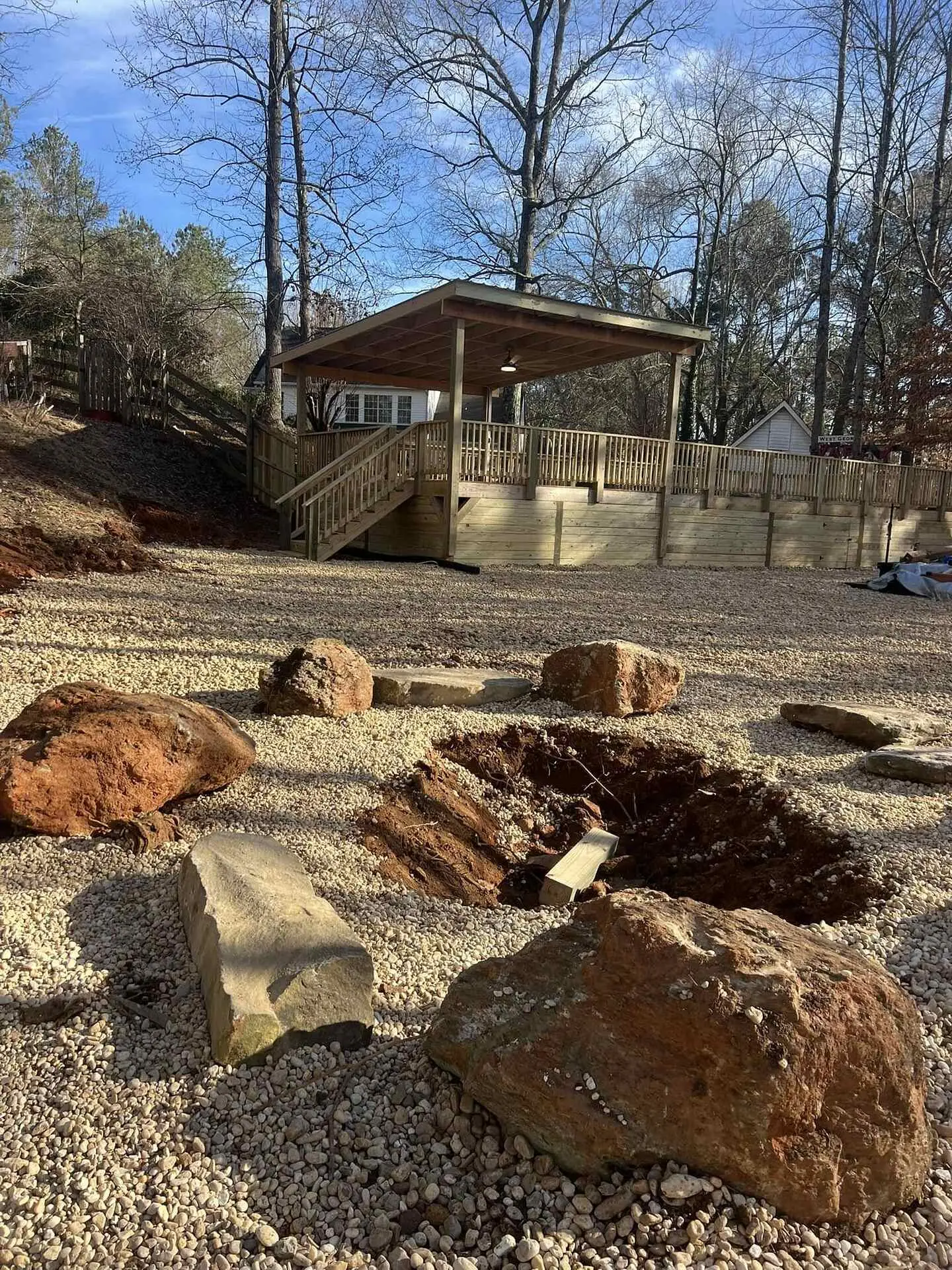 Elevated covered pavilion deck with timber walls enclosing the space beneath it, landscape boulders and gravel spread in front, an excavation still open in the gravel
