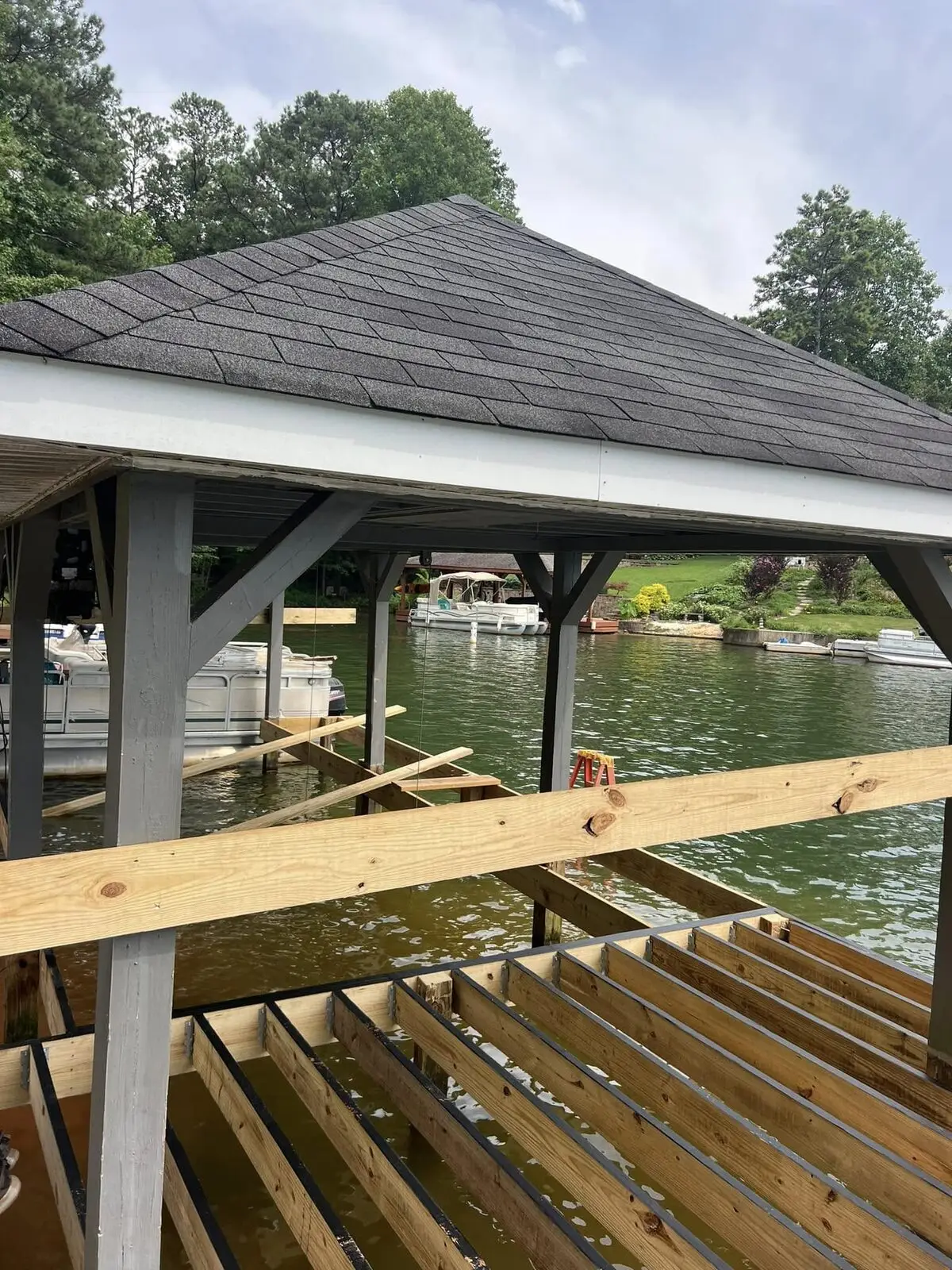 New pressure-treated dock joists set on galvanized hangers under a covered boathouse, lake beyond; tools and lumber staged on the deck, no crew member in frame
