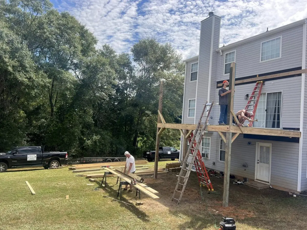 Crew framing an elevated second-story deck addition, ladders and lumber staged in the yard