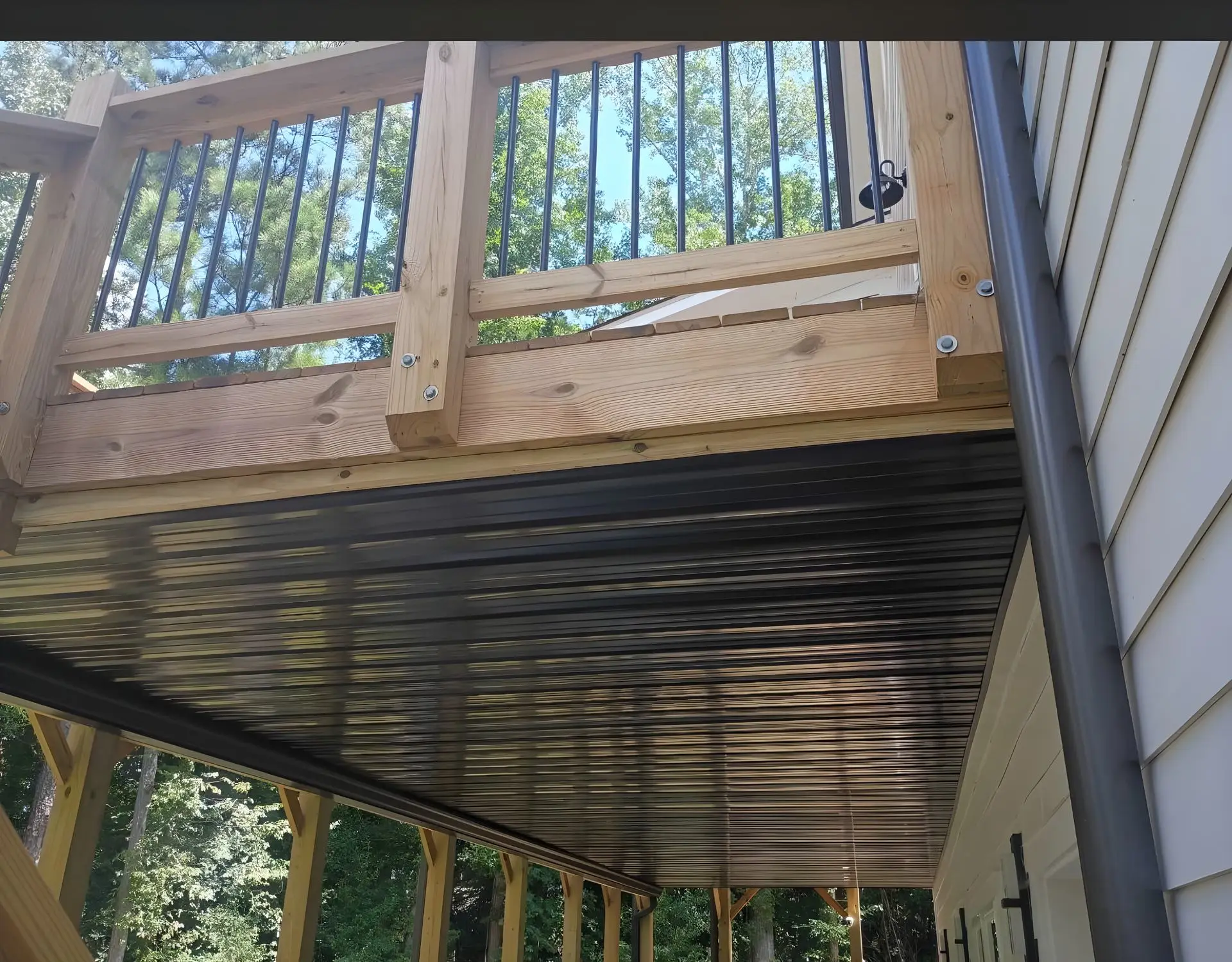 Underside view of an elevated deck looking up at a black corrugated metal ceiling panel and a wood rail with black metal balusters above, house siding alongside, trees in the background