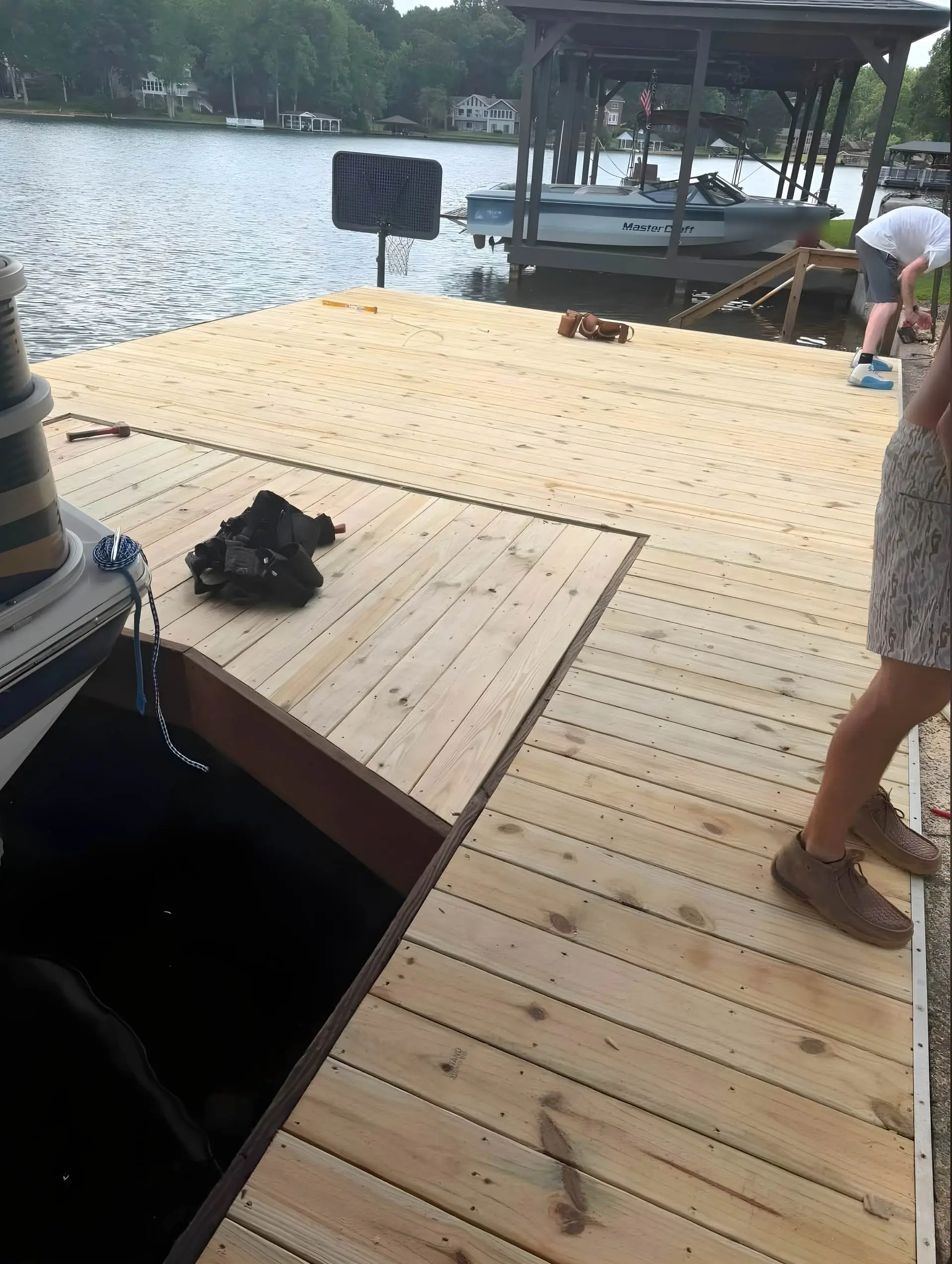 New dock decking installation with an open trapdoor/access hatch to the water below, crew member working near a covered boat lift with a docked ski boat, basketball hoop mounted at the dock edge.