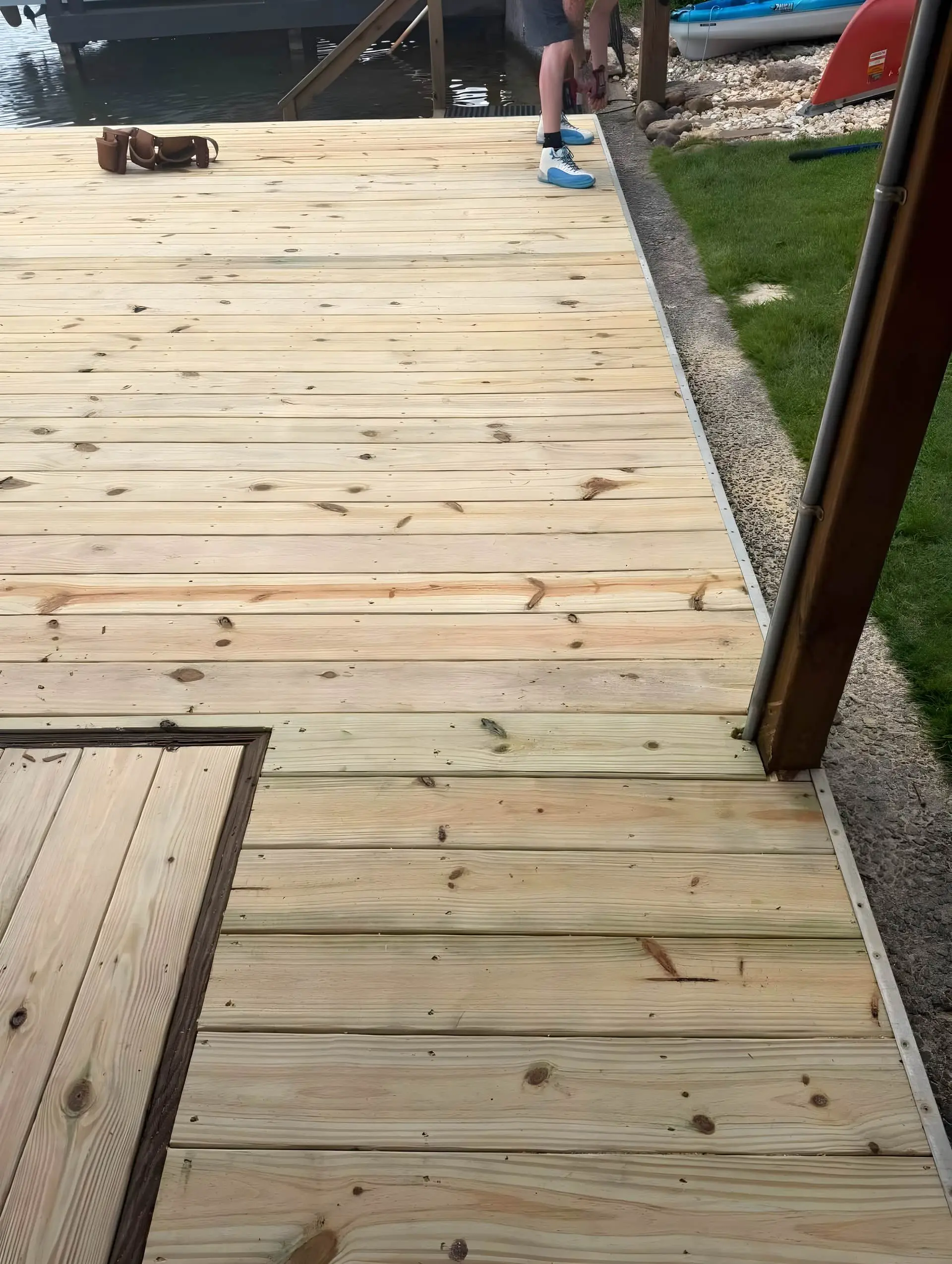 New pressure-treated dock decking boards freshly installed, no rail yet, boat ramp/steps visible at the dock edge, kayaks and grass beyond.