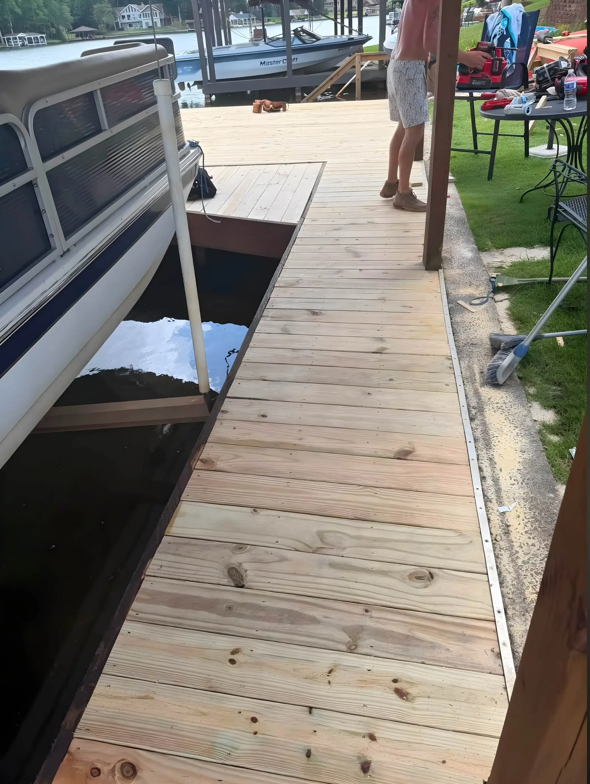 New dock decking installed with an open hatch section near the boat lift, pontoon boat and ski boat moored alongside, crew member standing at the far end near tools staged on a table