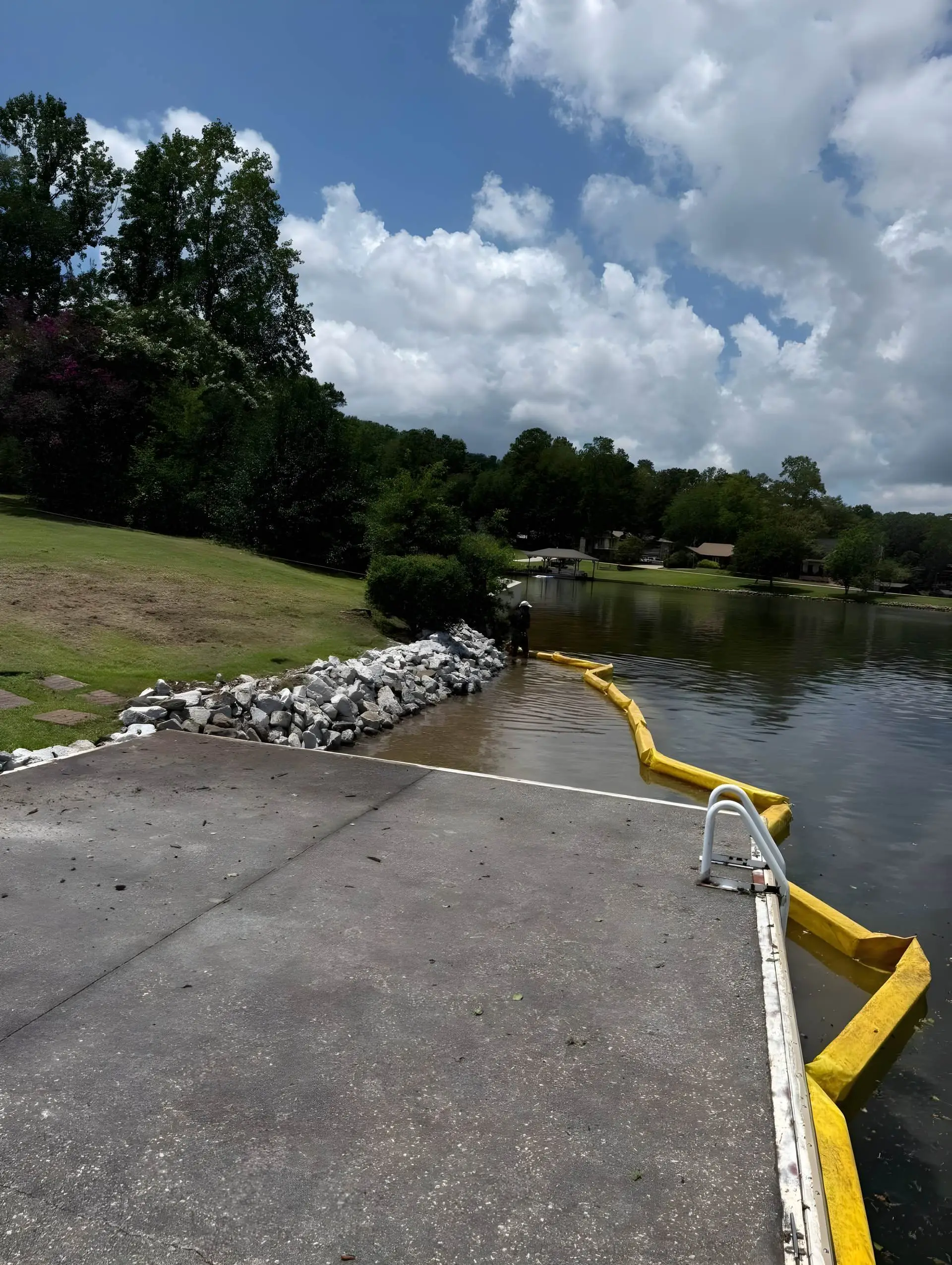 Worker placing riprap stone along a shoreline/seawall repair, a yellow silt curtain floating in the water to contain sediment.