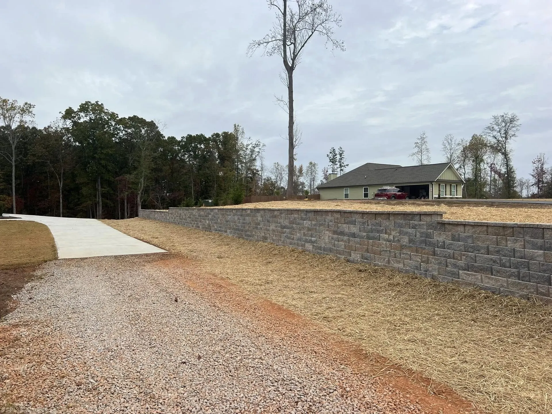 Long segmental block retaining wall holding a raised yard above a gravel drive, freshly graded and strawed