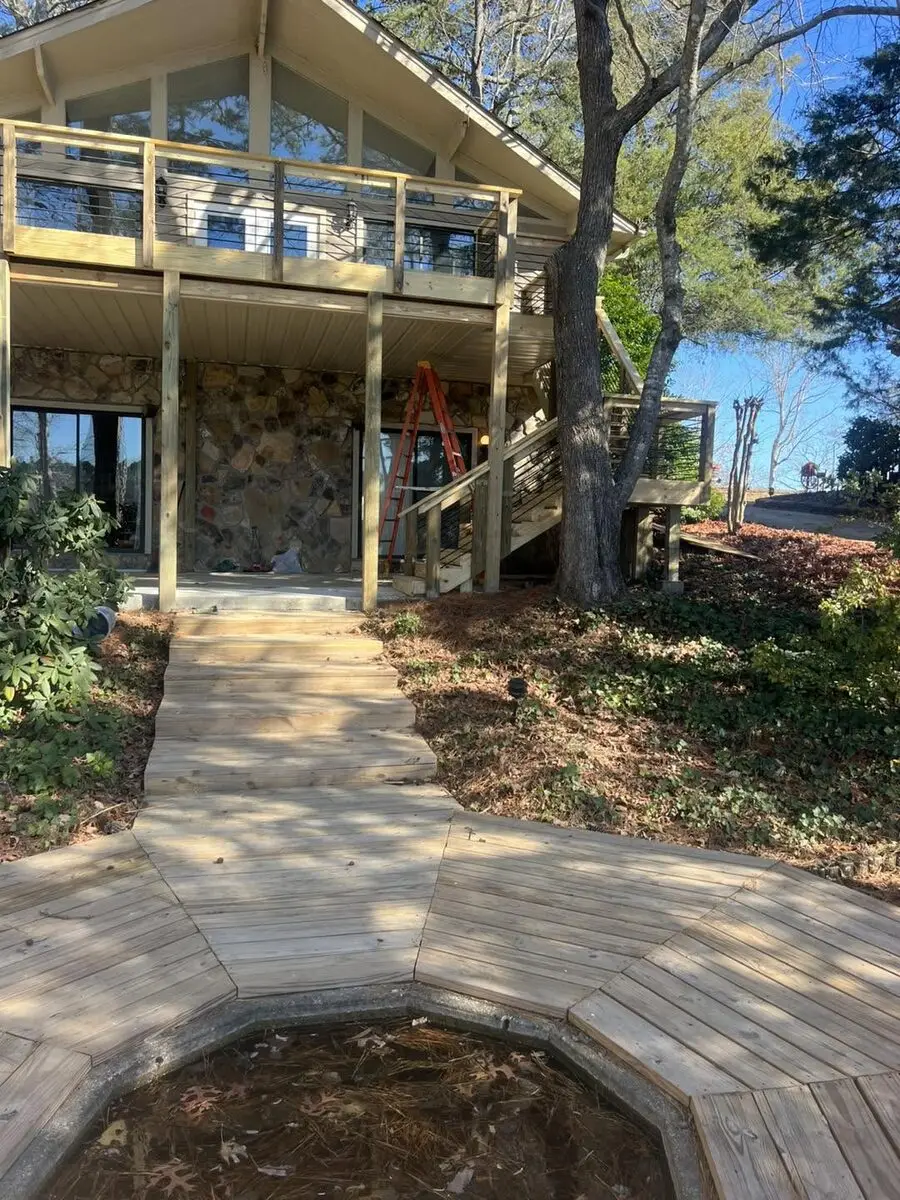Cable-rail deck above and a new wood boardwalk below at a chalet-style home, the boardwalk wrapping an empty garden pond basin, mid-build