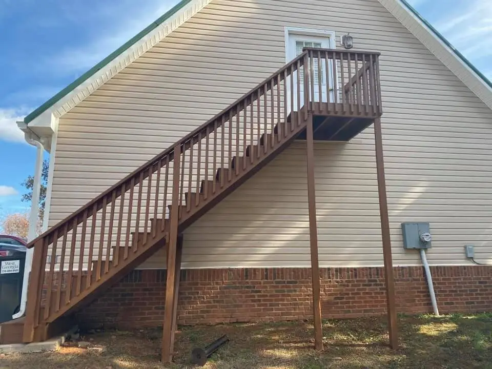 Two-story brown-stained deck staircase against tan siding, company-branded vehicle parked in the driveway